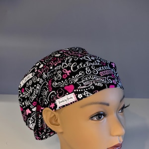 May include: A black and white patterned surgical scrub cap with pink accents. The cap features hearts, ribbons, and text including "Cure", "My Courage", and "Survivor". A small white label reads "Brands by Brooks".