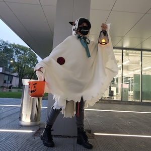 May include: A person dressed as a ghost for Halloween, wearing a white hooded costume with a ripped hem, holding a black lantern and a pumpkin-shaped bucket. The person is wearing a black face mask and a teal scarf.