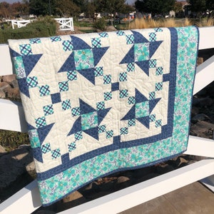 May include: A blue and white quilt with a star pattern. The quilt is made with a variety of fabrics, including gingham and floral prints. The quilt is hanging on a white fence.