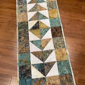 May include: A rectangular quilted table runner with a patchwork design. The runner features squares and triangles in shades of teal, brown, gold, and white. The edges are finished with a light blue border. The overall design is geometric.