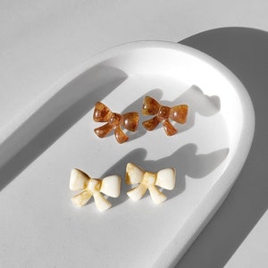 Peut inclure: Quatre boucles d'oreilles en forme de nœud en résine, deux brunes et deux blanches, présentées sur une assiette en céramique blanche.