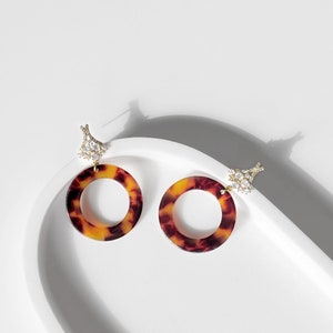 May include: A pair of gold tone earrings with a tortoise shell design. The earrings feature a round hoop with a clear crystal cluster at the top.