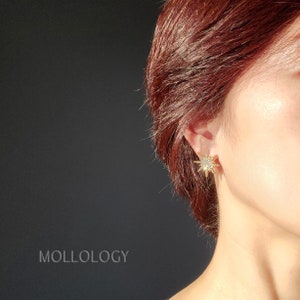 May include: Gold star-shaped earrings with clear rhinestones. The earrings are worn in the ear of a person with red hair.