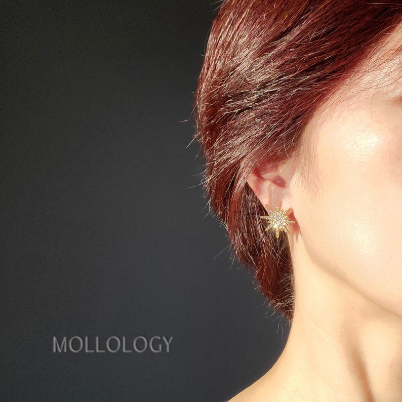 May include: Gold star-shaped earrings with clear rhinestones. The earrings are worn in the ear of a person with red hair.