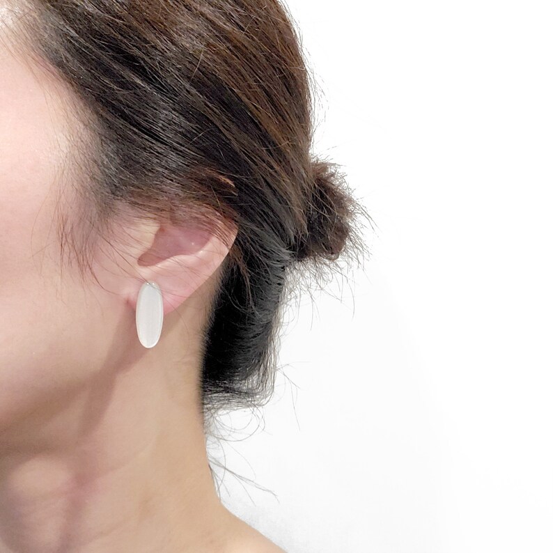 May include: A pair of oval-shaped earrings with a white, opaque finish. The earrings are worn by a person with brown hair.