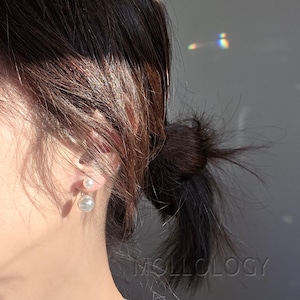 Puede incluir: Pendientes de perlas blancas. Cada pendiente tiene una pequeña perla como botón y una perla redonda más grande debajo. Cabello oscuro recogido en un moño. La palabra "MOLLOLOGY" es visible en la parte inferior derecha.