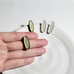 May include: Three oval-shaped earrings, two are white and one is green. The earrings are on a white surface.