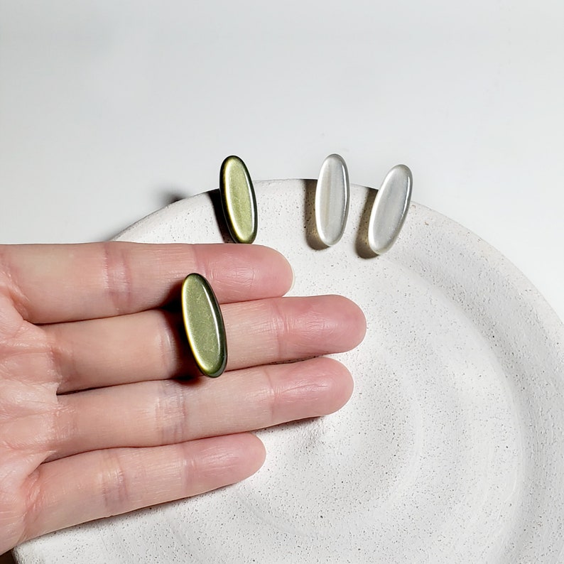May include: Three oval-shaped earrings, two are white and one is green. The earrings are on a white surface.