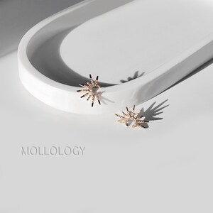 May include: A pair of gold starburst earrings with clear crystal accents. The earrings are on a white surface.