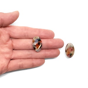 Puede incluir: Un par de pendientes ovalados con un patr&oacute;n abstracto colorido. Los pendientes est&aacute;n hechos de un material transl&uacute;cido y tienen un fondo blanco con salpicaduras de rojo, naranja, azul y verde.