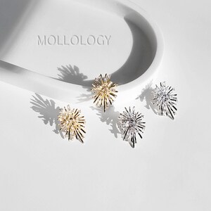 May include: Three pairs of starburst-shaped earrings in gold, silver, and rose gold. The earrings are on a white surface with the word "MOLLOLOGY" printed on the surface.