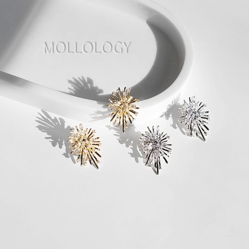 May include: Three pairs of starburst-shaped earrings in gold, silver, and rose gold. The earrings are on a white surface with the word "MOLLOLOGY" printed on the surface.