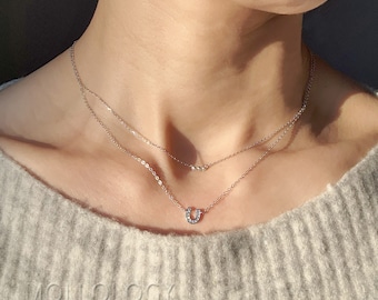 Puede incluir: Un delicado collar de oro con un colgante de herradura incrustado con pequeñas piedras transparentes. El collar está superpuesto con una cadena más fina con un pequeño acento de piedra transparente.