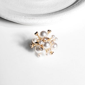 Könnte beinhalten: Ein goldener Ring mit einem Cluster aus weißen Perlen und klaren Strasssteinen. Der Ring ist so gestaltet, dass er wie ein Lichtschwall aussieht.