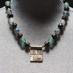 May include: A silver pendant necklace with a geometric design hangs from a strand of frosted glass beads with bronze accents.