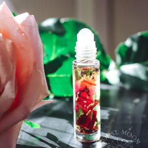 Könnte beinhalten: Eine transparente Glas-Rollerflasche gefüllt mit einer goldenen Flüssigkeit und getrockneten roten Blütenblättern und grünen Blättern. Eine rosa Rose und grünes Laub befinden sich im Hintergrund. Die Flasche hat einen weißen Deckel. Das Opal Moon Apothecary-Logo ist sichtbar.