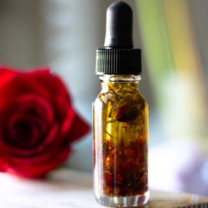 May include: A small clear glass dropper bottle filled with a golden-colored oil containing visible dried botanical pieces and small red petals. The bottle is topped with a black ribbed cap and a black rubber dropper bulb.