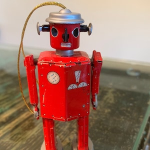 May include: A red robot toy ornament with a silver head and a gold string. The robot has a clock face and gauges on its chest.