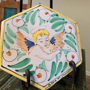 May include: A hexagonal ceramic tile with a hand-painted design of a cherub with wings, a halo, and a flower. The tile is decorated with green, yellow, and orange swirls and the text "D. Lucca all' Cavaliere".