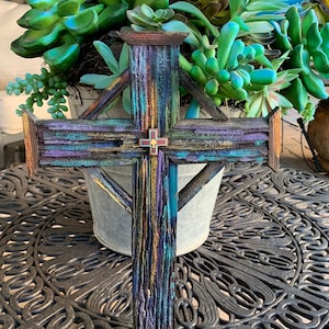 May include: A wooden cross with a distressed, multi-colored paint finish. The cross is painted in shades of blue, purple, green, and yellow. A small red cross is centered on the front of the cross.