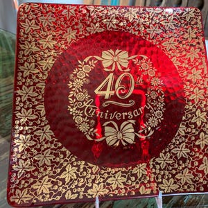 May include: A square, red glass plate with a gold floral pattern and the text "40th Anniversary" in a central circle. The plate has a textured surface and is supported by a white stand.