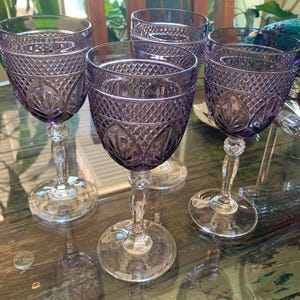 May include: Four amethyst-colored wine glasses with diamond-cut patterns. The glasses have a rounded bowl shape, a clear stem, and a circular base. The glasses are arranged on a reflective surface.