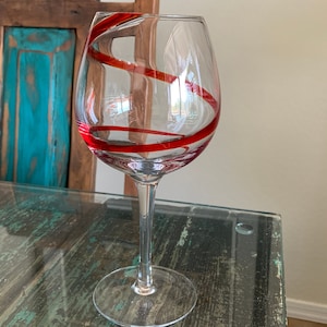 May include: A clear glass wine glass with a red swirl design. The glass is sitting on a wooden table.