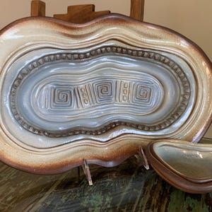 May include: A ceramic serving platter and small dish. The platter is oval with a light beige exterior and a blue-gray interior with spiral and linear designs. The dish is a smaller version of the platter, with a similar color scheme.