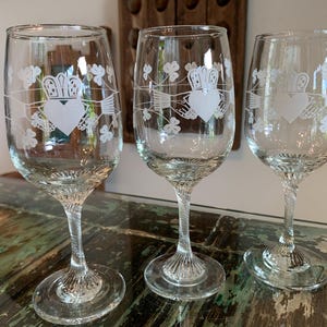 May include: Three clear wine glasses with a Claddagh design etched on the front. The design includes a heart, crown, and hands, along with shamrocks. The glasses have a twisted stem and a round base, perfect for serving wine.