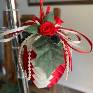 May include: A handmade Christmas ornament shaped like a pine cone, decorated with red and white ribbon, green fabric leaves, a red fabric rose, and white pearl beads.