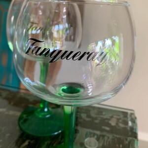 May include: Clear glass gin glasses with green stems. The word "Tanqueray" is printed in black script on the front of the glass. The glasses are designed for cocktails and other beverages.