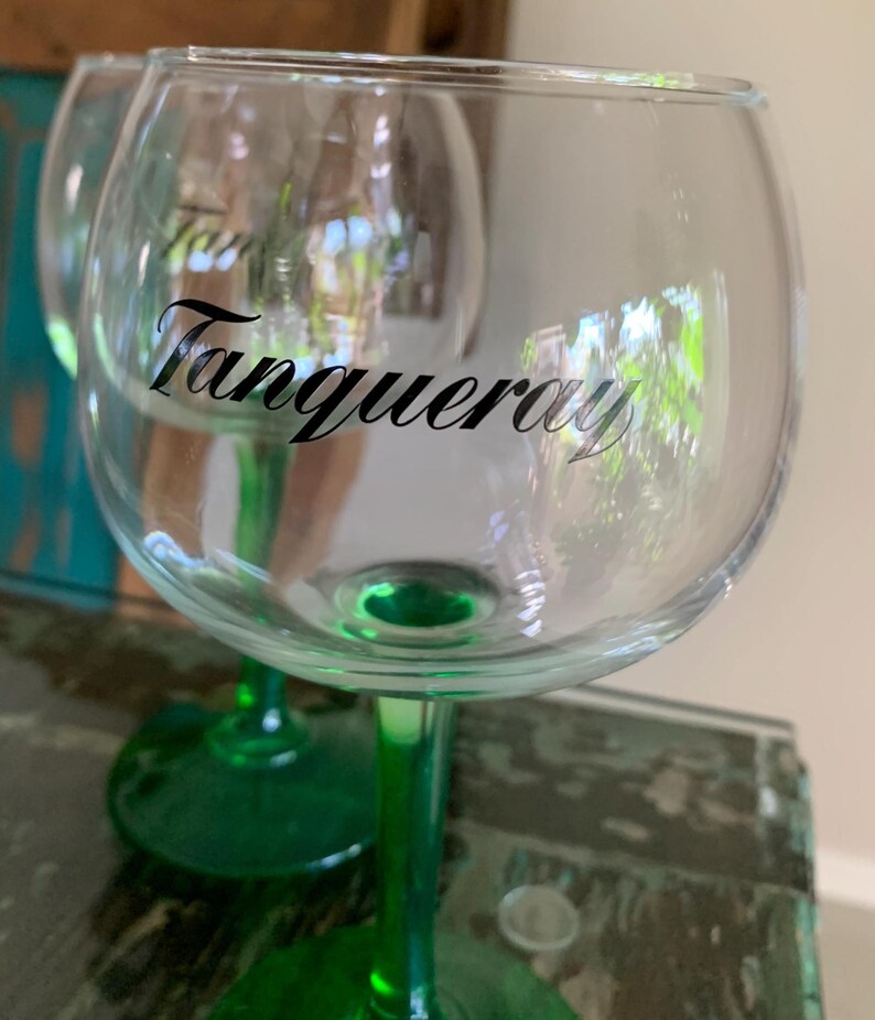 May include: Clear glass gin glasses with green stems. The word "Tanqueray" is printed in black script on the front of the glass. The glasses are designed for cocktails and other beverages.