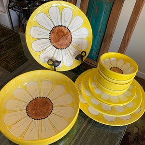 May include: A set of yellow and white ceramic dinnerware with a daisy design. The set includes a large platter, a smaller plate, and four bowls.