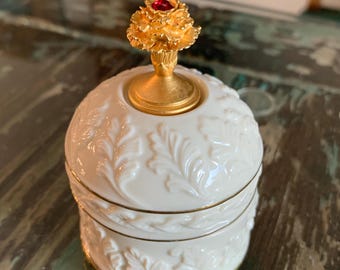 以下が含まれることがあります： 白い丸い装飾ジャー。金色の蓋には花の模様と赤い宝石が付いています。ジャーには、詳細なエンボス加工の葉のパターンと金のトリムがあります。