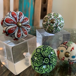 May include: Four ceramic drawer knobs with floral designs. The knobs are white with red, green, and blue floral patterns. The knobs are displayed on clear acrylic blocks.