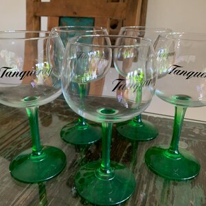 May include: Six clear, stemmed gin glasses with green bases and stems. The word "Tanqueray" is printed in black on the side of each glass. The glasses are arranged on a glass-topped table.