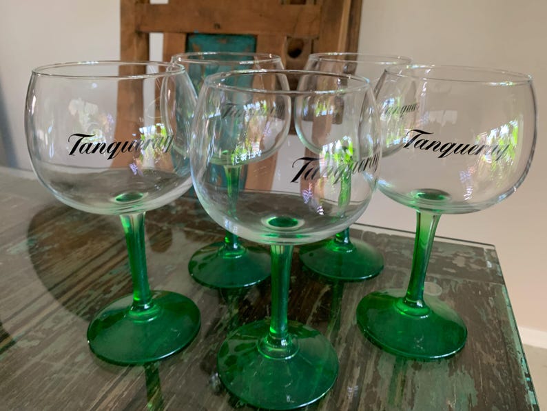 May include: Six clear, stemmed gin glasses with green bases and stems. The word "Tanqueray" is printed in black on the side of each glass. The glasses are arranged on a glass-topped table.
