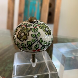 May include: A white ceramic knob with a floral design in shades of green, yellow, and red. The knob is round and has a brass screw.