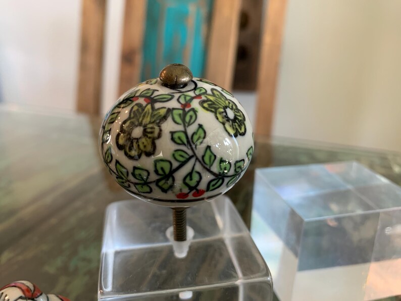 May include: A white ceramic knob with a floral design in shades of green, yellow, and red. The knob is round and has a brass screw.