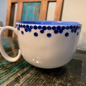 May include: A white ceramic mug with a blue honeycomb pattern around the rim. The mug has a rounded shape and a handle.
