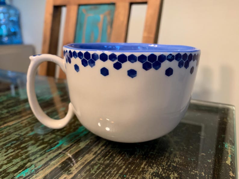 May include: A white ceramic mug with a blue honeycomb pattern around the rim. The mug has a rounded shape and a handle.