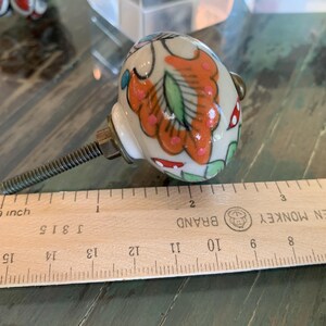 May include: A white ceramic knob with a colorful floral design in shades of orange, pink, green, and red. The knob is attached to a silver screw and is measured against a wooden ruler labeled "GOLDEN MONKEY BRAND".