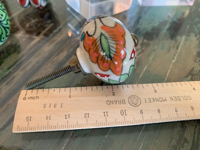 May include: A white ceramic knob with a colorful floral design in shades of orange, pink, green, and red. The knob is attached to a silver screw and is measured against a wooden ruler labeled "GOLDEN MONKEY BRAND".