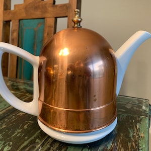 May include: A white teapot with a copper lid and a brass knob. The teapot is sitting on a wooden table.