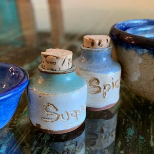 May include: A collection of handmade ceramic kitchenware. Includes a small blue bowl, two small jars labeled "Sugar" and "Spice" with cork stoppers, and a nested bowl set with a berry design. The pottery has a rustic, handcrafted appearance.