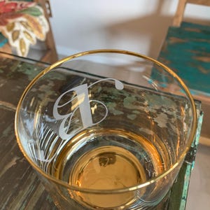 May include: A clear glass tumbler with a gold rim and base. The glass is etched with a stylized letter "B". The glass is sitting on a distressed wooden surface.