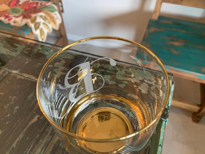 May include: A clear glass tumbler with a gold rim and base. The glass is etched with a stylized letter "B". The glass is sitting on a distressed wooden surface.