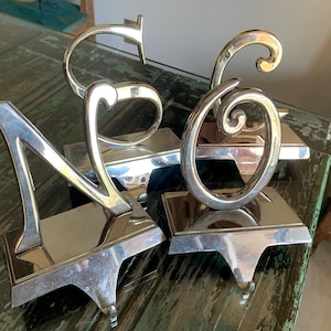 May include: Four silver metal stocking holders spelling the word "NOEL". Each letter is attached to a rectangular base with a hook. The letters have a shiny, reflective surface and are displayed on a green and brown surface.
