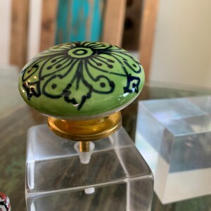 May include: A green ceramic knob with a black and white floral design, mounted on a clear acrylic base.