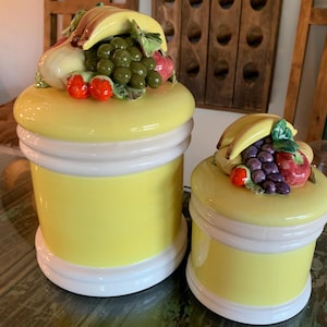 May include: Two yellow ceramic canisters with white bands at the base and lid. Each lid features a sculpted, hand-painted arrangement of fruit including bananas, grapes, and apples. The canisters have a smooth, glossy finish and cylindrical bodies.